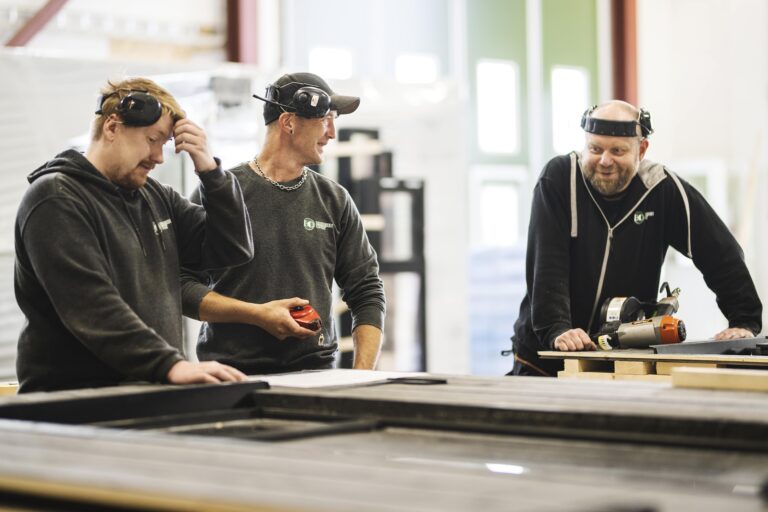 bildet viser tre menn som smiler og snakker sammen i en produksjonshall